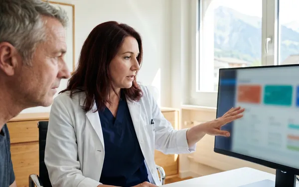 Ärztin in Schweizer Praxis berät einen Patienten zur medizinischen Cannabis-Therapie vor einem Computerbildschirm mit Behandlungsdaten